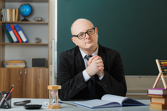 Middle-aged man with glasses sitting at desk in classroom, appearing like an obnoxious dinner guest husband dreadful. Middle-aged man with glasses sitting at desk in classroom, appearing like an obnoxious dinner guest husband dreadful.