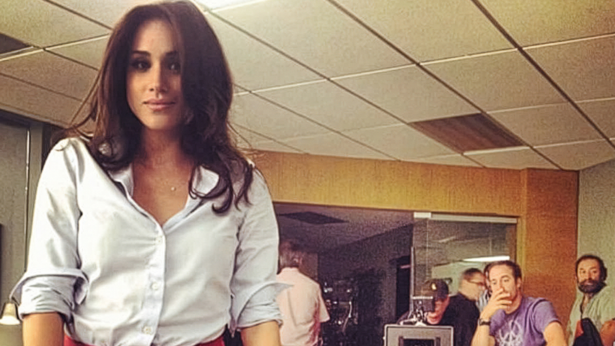 Meghan Markle standing indoors wearing a white blouse, with a casual office background, related to net worth figures 2025.
