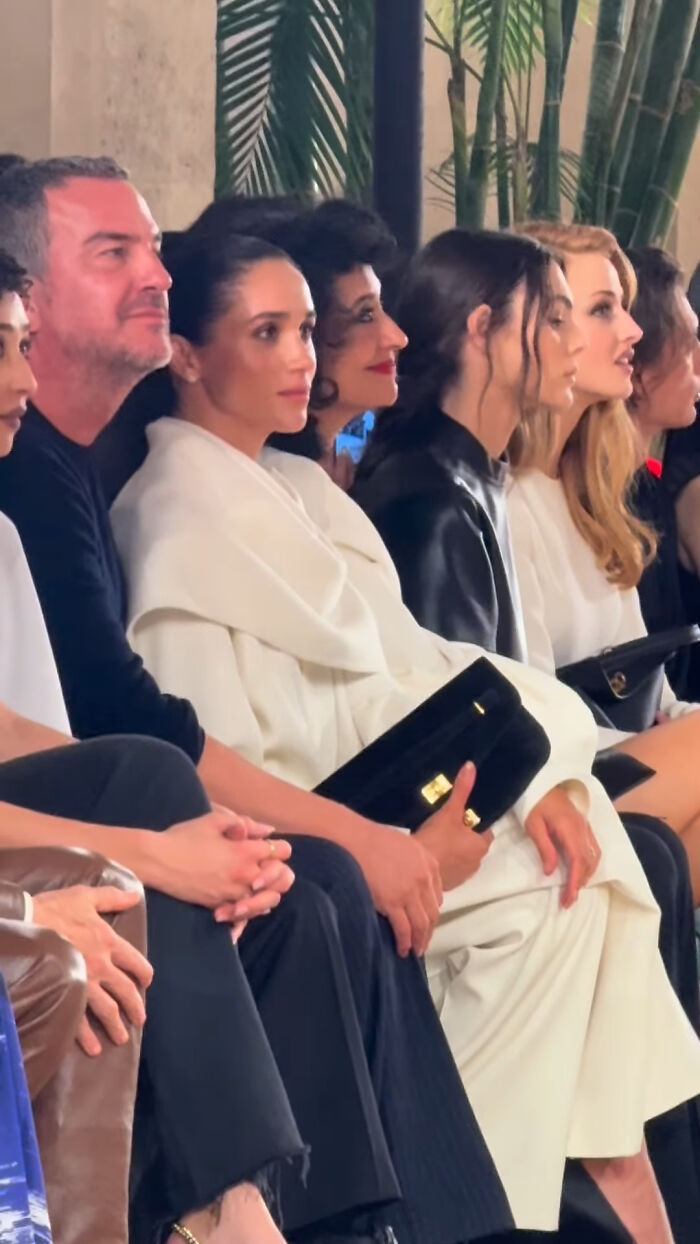 Meghan Markle sitting front row at Paris Fashion Week, dressed in white, holding a black clutch amid other fashion attendees. Meghan Markle sitting front row at Paris Fashion Week, dressed in white, holding a black clutch amid other fashion attendees.