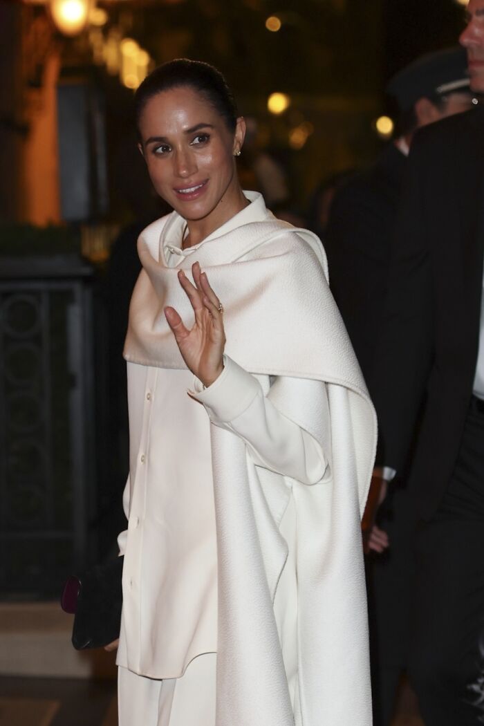 Meghan Markle smiling and waving at Paris Fashion Week, wearing an elegant white outfit during a nighttime event. Meghan Markle smiling and waving at Paris Fashion Week, wearing an elegant white outfit during a nighttime event.