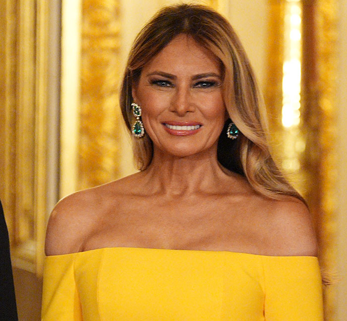 Melania Trump wearing a yellow off-shoulder dress and emerald earrings at a White House event with golden decor background Melania Trump wearing a yellow off-shoulder dress and emerald earrings at a White House event with golden decor background