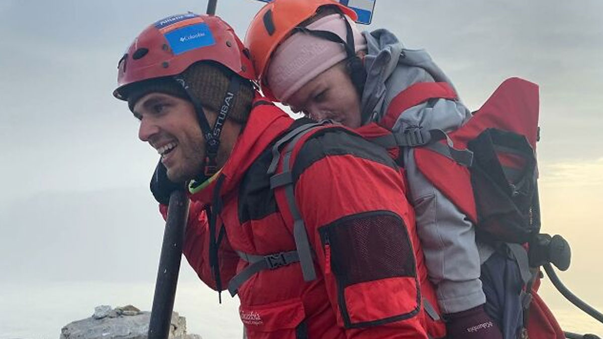 Man wearing a red helmet and jacket carrying a person on his back, showing the good in this world through kindness.