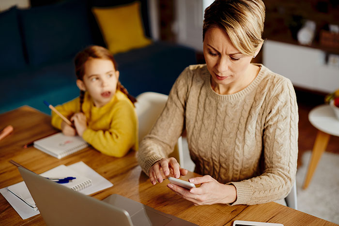 Mother working on smartphone at table while daughter in yellow sweater looks on, highlighting toxic in-laws conflict and newborn issues. Mother working on smartphone at table while daughter in yellow sweater looks on, highlighting toxic in-laws conflict and newborn issues.