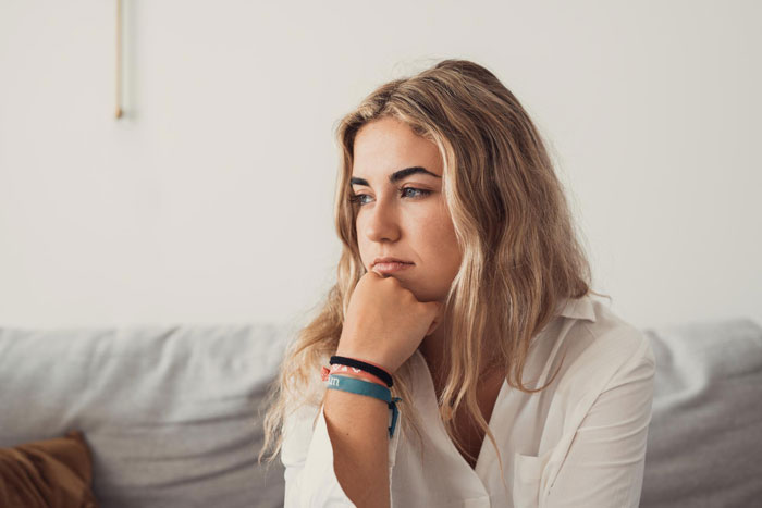 Young woman with blonde hair and white shirt sitting thoughtfully on a couch reflecting on rude MIL behavior issues. Young woman with blonde hair and white shirt sitting thoughtfully on a couch reflecting on rude MIL behavior issues.