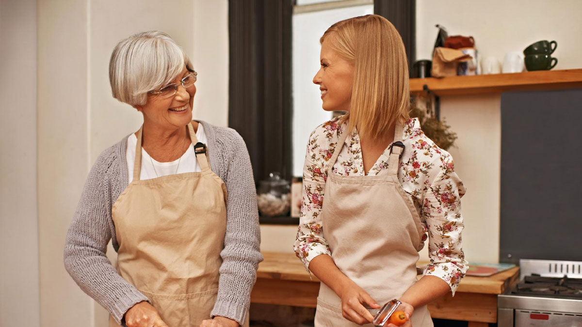 Older woman and younger woman wearing aprons smiling at each other while preparing food in a kitchen setting