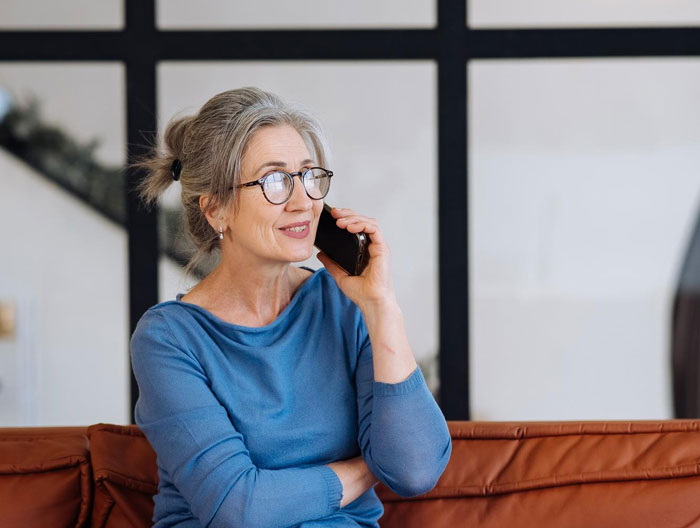 Middle-aged woman wearing glasses sitting on couch, talking on phone, representing DIL receiving mysterious nonstop calls. Middle-aged woman wearing glasses sitting on couch, talking on phone, representing DIL receiving mysterious nonstop calls.