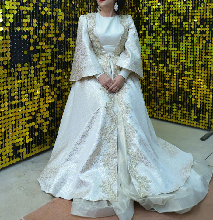 Woman in ornate white wedding dress with gold embroidery standing against a sequin gold background at a wedding event. Woman in ornate white wedding dress with gold embroidery standing against a sequin gold background at a wedding event.
