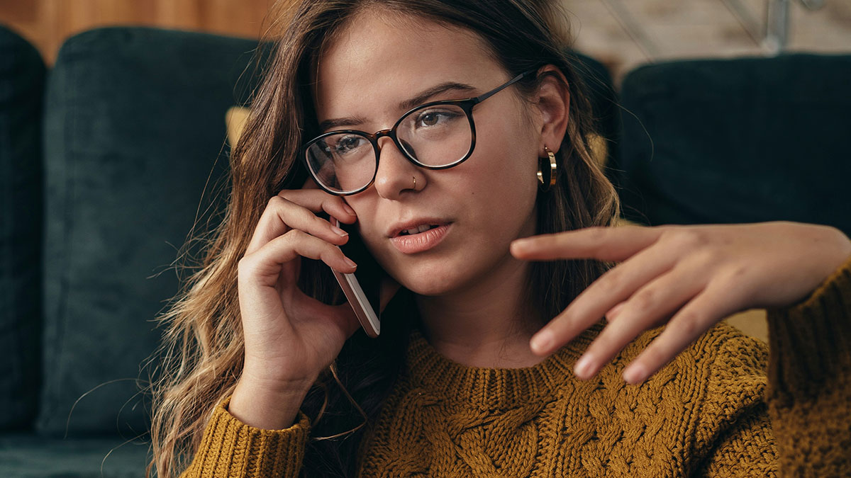 Young woman in glasses and mustard sweater making a phone call, showing a socially awkward situation expression.