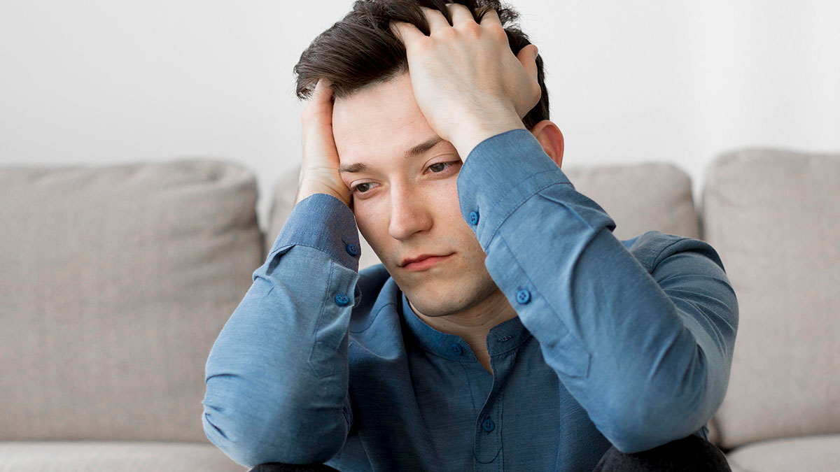 Young man in a blue shirt sitting on a couch, holding his head in frustration, reflecting on bio mom abandoning him.