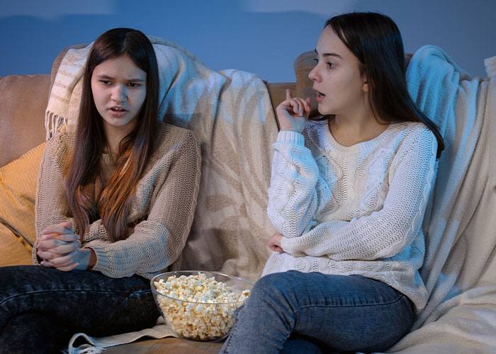 Two teenage daughters sitting on a couch with popcorn, showing tension while mom admits she likes one daughter more than the other. Two teenage daughters sitting on a couch with popcorn, showing tension while mom admits she likes one daughter more than the other.