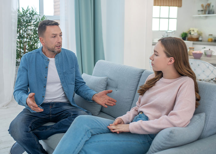 Man and teenage daughter having a serious conversation on a couch, reflecting struggles of mom admitting favoritism between daughters. Man and teenage daughter having a serious conversation on a couch, reflecting struggles of mom admitting favoritism between daughters.