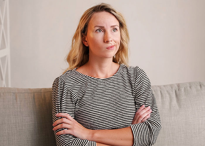 A mom sitting on a couch with crossed arms, showing a hesitant expression about liking one daughter more than the other. A mom sitting on a couch with crossed arms, showing a hesitant expression about liking one daughter more than the other.