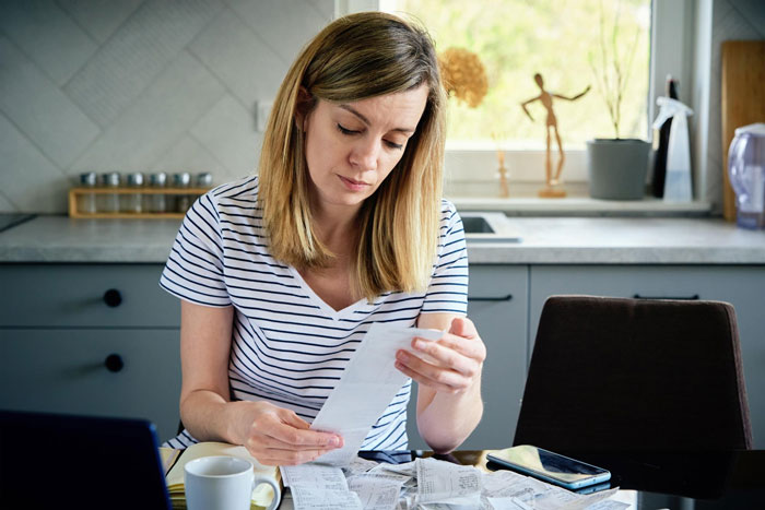 Woman venting about useless bro, looking frustrated while reviewing receipts at kitchen table with phone and laptop nearby Woman venting about useless bro, looking frustrated while reviewing receipts at kitchen table with phone and laptop nearby