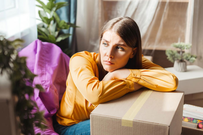 Young woman in a yellow shirt looking upset while leaning on a cardboard box, venting about her useless brother. Young woman in a yellow shirt looking upset while leaning on a cardboard box, venting about her useless brother.