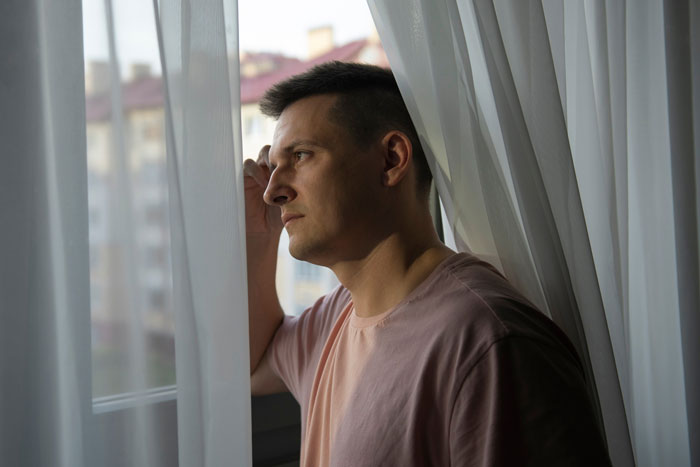 Man looking thoughtfully out window, concerned about male Karen blocking neighbor's driveway repeatedly