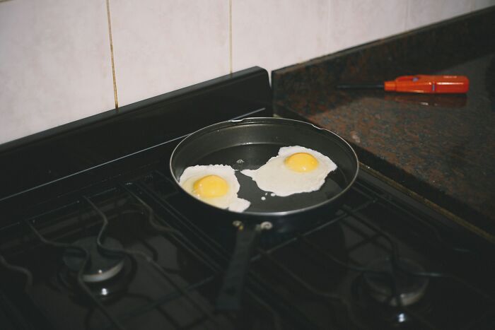 Two eggs frying in a black pan on a gas stove in a kitchen with a dark countertop and wall tiles nearby.