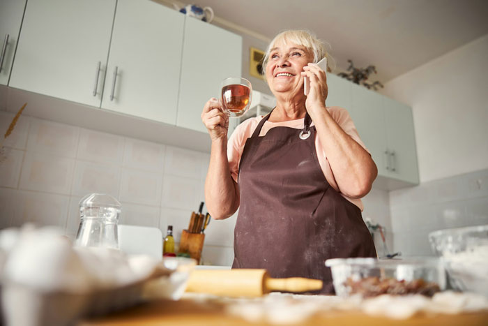 Elderly woman in kitchen wearing apron, holding tea and phone, representing nosy mother with ancestry tests theme. Elderly woman in kitchen wearing apron, holding tea and phone, representing nosy mother with ancestry tests theme.