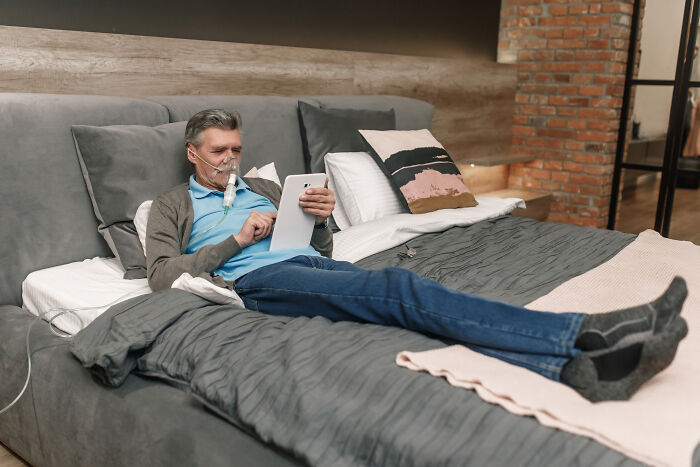 Older man using oxygen therapy at home, reading from a tablet while resting in bed, showing patient health monitoring.