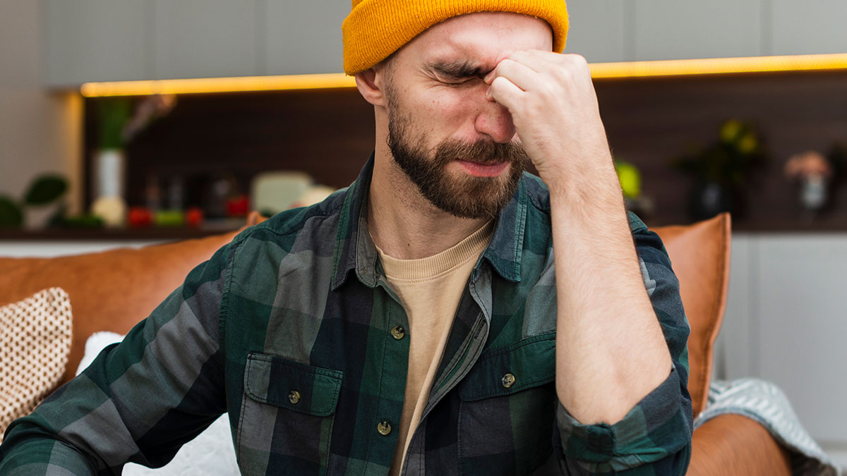 Man wearing beanie and plaid shirt, pinching bridge of nose in frustration, reflecting arrogant professor at party misery.