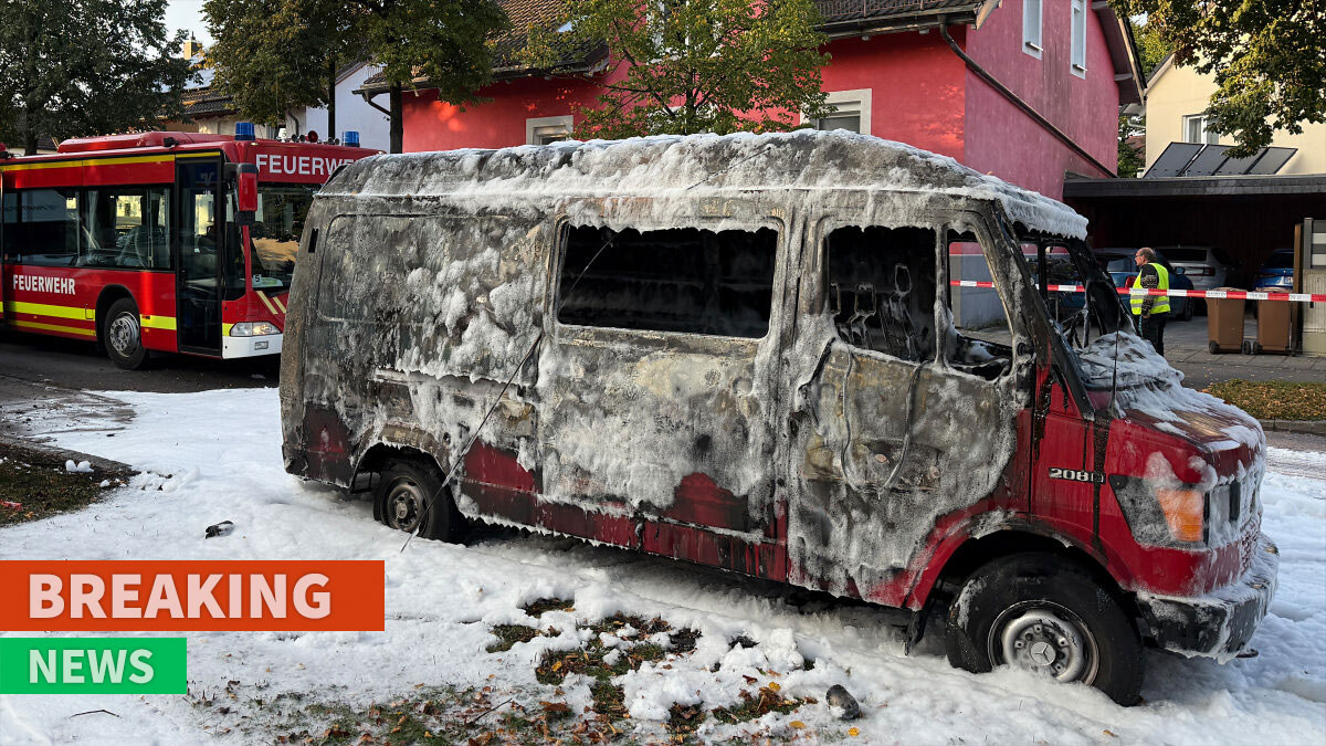 Burned vehicle covered in foam at explosion site in Munich with fire truck and emergency responders nearby.