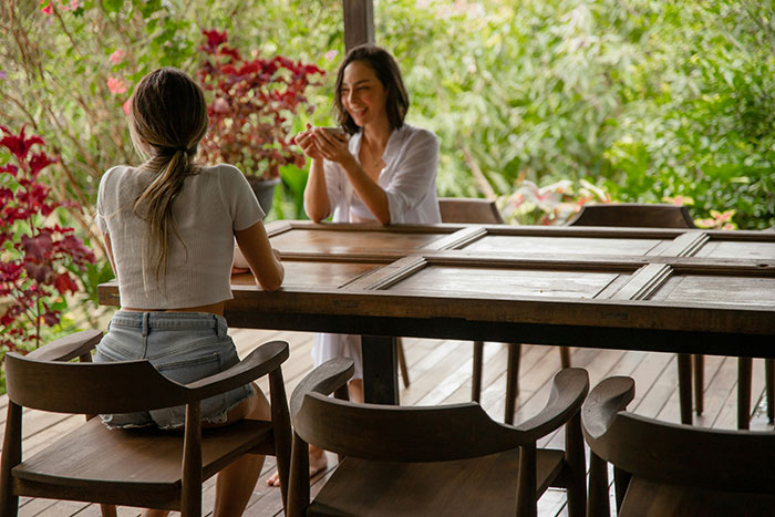 Two women sitting at a wooden table outdoors, one smiling while holding a cup, depicting a fiancée and her friend. Two women sitting at a wooden table outdoors, one smiling while holding a cup, depicting a fiancée and her friend.