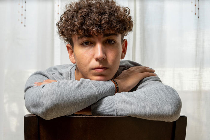 Teenage boy with curly hair sitting with arms crossed on chair, reflecting on toxic parents favoring golden child son. Teenage boy with curly hair sitting with arms crossed on chair, reflecting on toxic parents favoring golden child son.