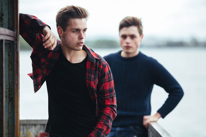 Two young men outdoors by water, one looking upset and distant, depicting tension between favored golden child and younger son. Two young men outdoors by water, one looking upset and distant, depicting tension between favored golden child and younger son.