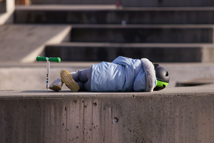 Teen wearing helmet and coat lying alone on concrete ledge next to a scooter in an empty urban area, symbolizing parents kicked out teen. Teen wearing helmet and coat lying alone on concrete ledge next to a scooter in an empty urban area, symbolizing parents kicked out teen.