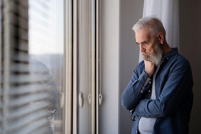 Elderly man with gray hair looking out window thoughtfully, illustrating themes of disabled brother care and family boundaries. Elderly man with gray hair looking out window thoughtfully, illustrating themes of disabled brother care and family boundaries.