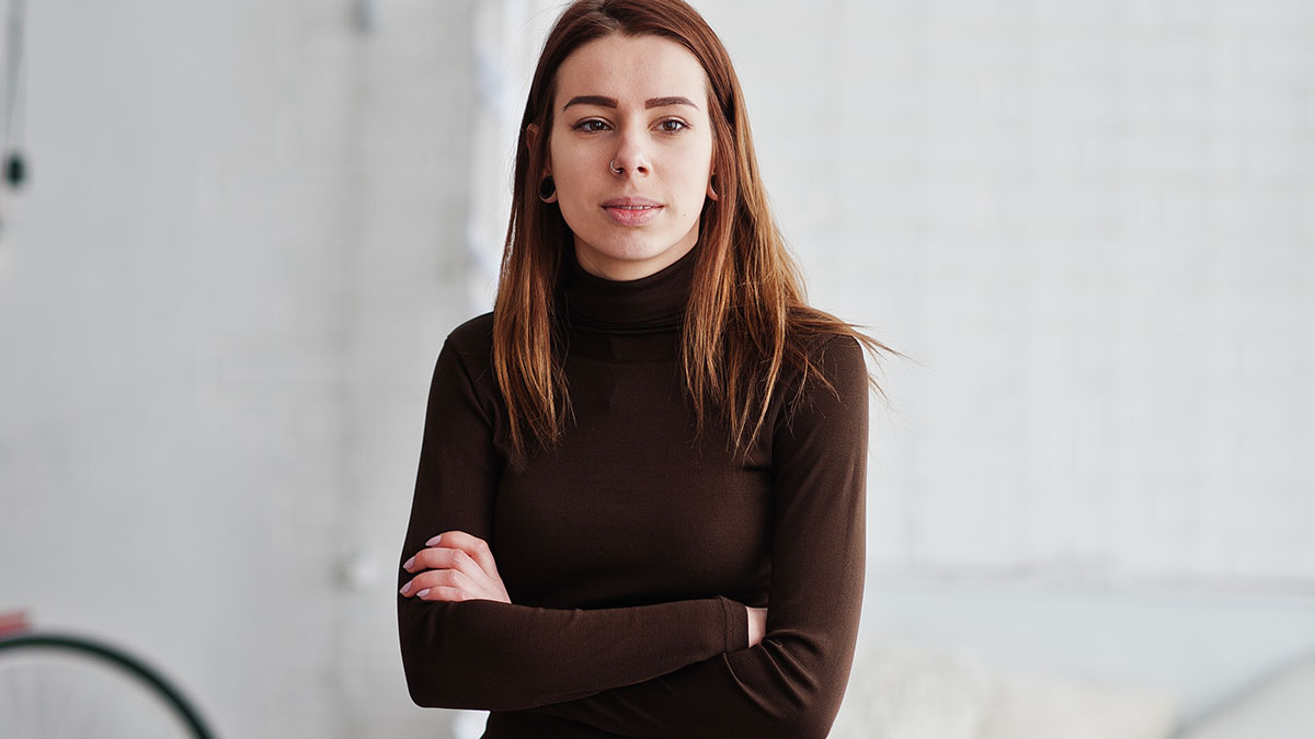 Young woman setting a boundary about disabled brotheru2019s care, standing with arms crossed in a neutral indoor space.