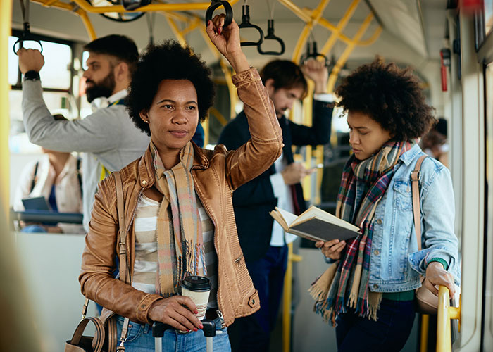 Woman standing on crowded bus holding handle and coffee while another passenger reads, illustrating speakerphone user stories. Woman standing on crowded bus holding handle and coffee while another passenger reads, illustrating speakerphone user stories.