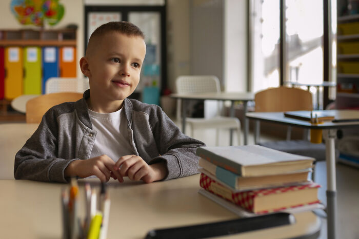 Young boy sitting at classroom desk with books, reflecting on childhood moments they still cringe about today.