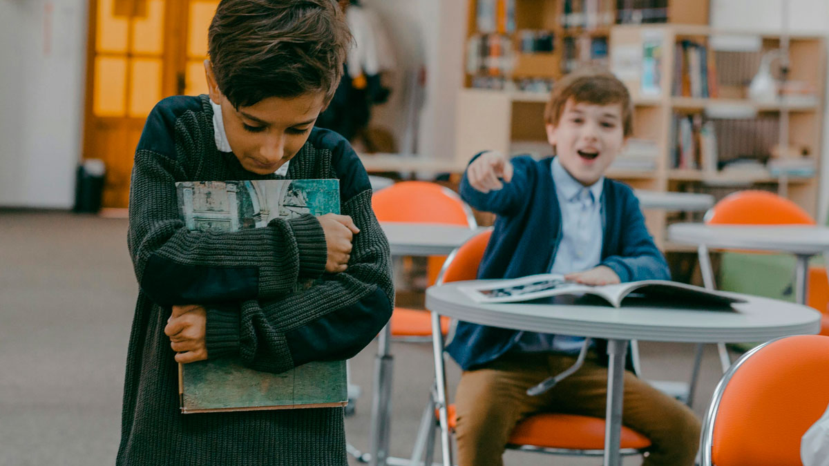 Two children in a classroom, one cringing while holding a book and the other pointing and laughing, showing childhood cringe moments.
