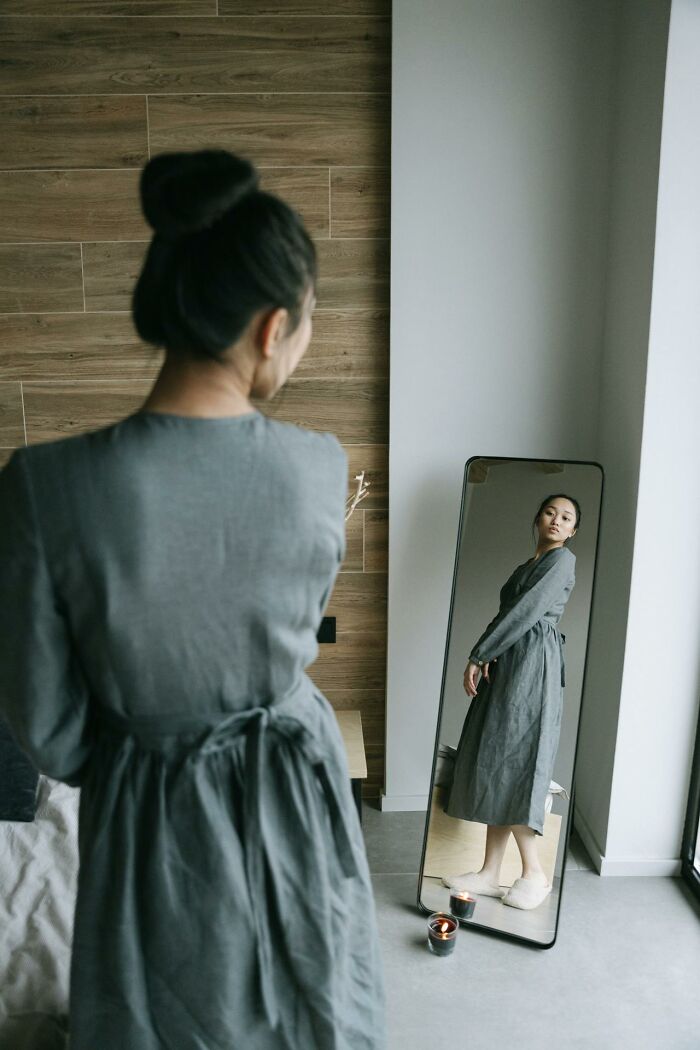 Woman in a gray dress looking at herself in a mirror in a modern room, reflecting signs someone is having an affair.