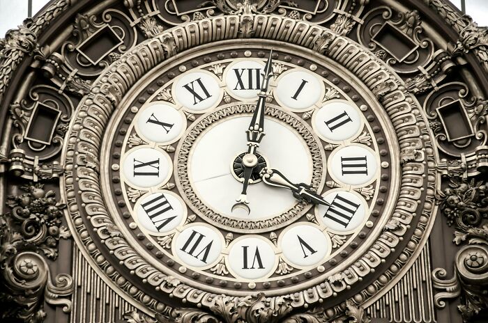 Ornate antique clock face with Roman numerals surrounded by intricate decorative carvings on a historical building facade. Ornate antique clock face with Roman numerals surrounded by intricate decorative carvings on a historical building facade.