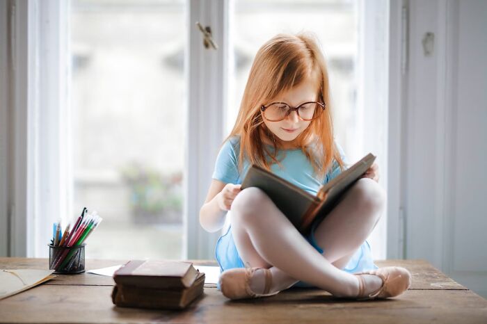 Young girl wearing glasses reading a book indoors, capturing a moment of focus related to disturbing things told to kids.