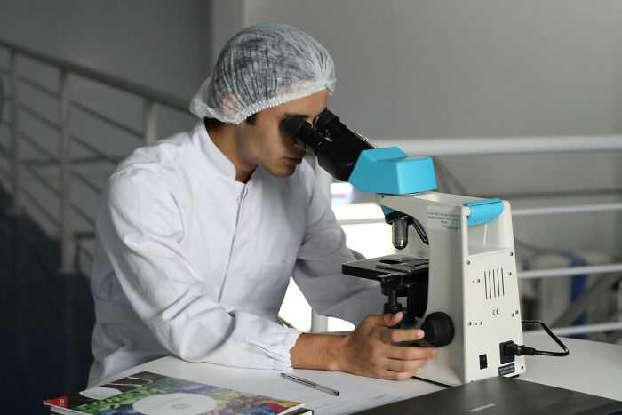 Scientist in a lab coat and hairnet examining samples under a microscope sharing insights on potentially dangerous situations.