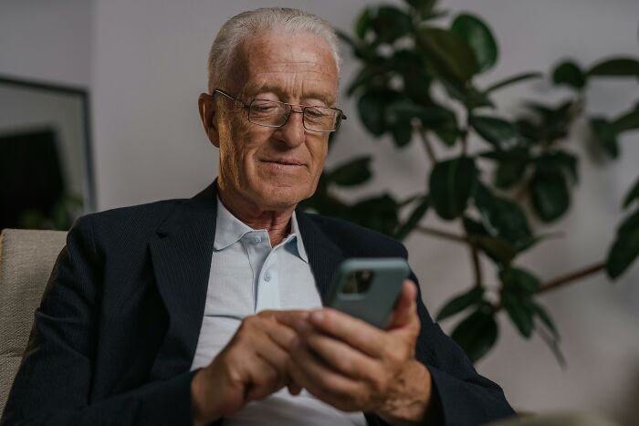 Older man wearing glasses, using smartphone indoors, illustrating the concept of genius life hacks that add extra work.