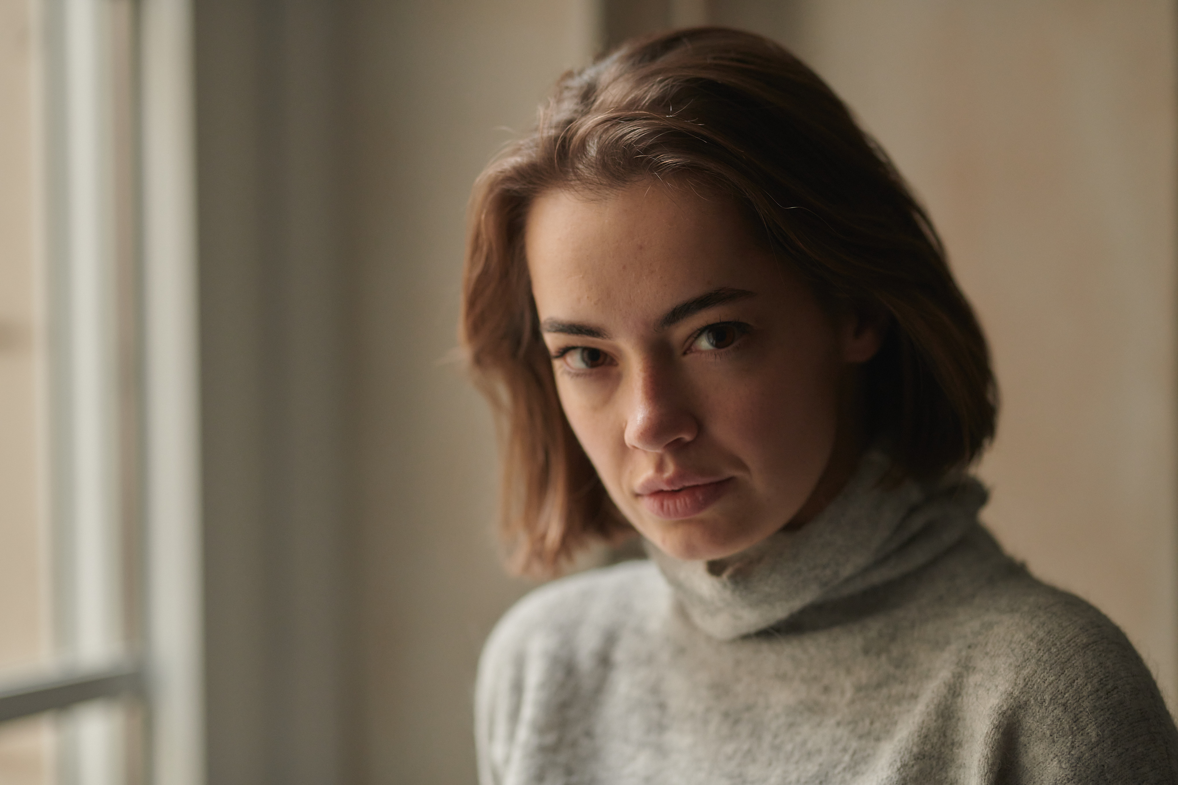 Young woman with short brown hair and gray turtleneck sweater looking serious by a window indoors. Young woman with short brown hair and gray turtleneck sweater looking serious by a window indoors.