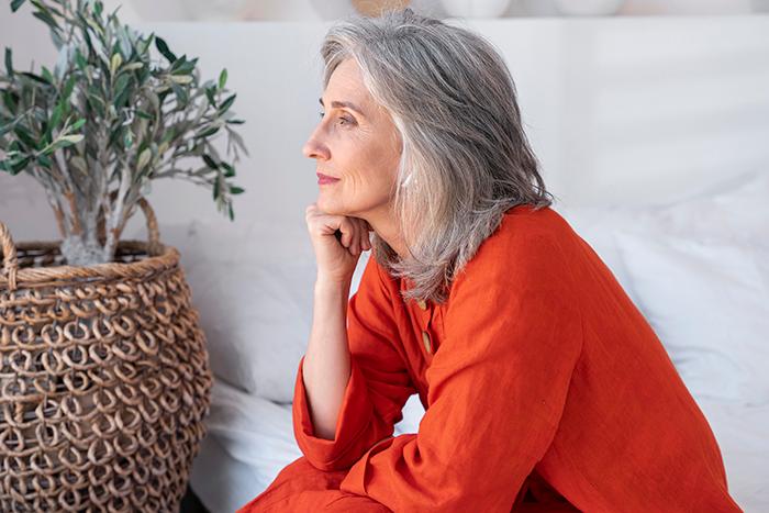 Middle-aged woman in orange dress sitting thoughtfully on bed near plant, reflecting on stepdaughters disown stepmother drama. Middle-aged woman in orange dress sitting thoughtfully on bed near plant, reflecting on stepdaughters disown stepmother drama.