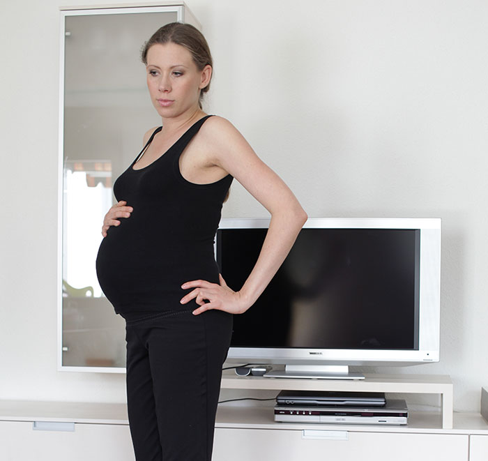 Pregnant woman standing with hand on belly and hip, looking concerned in a modern living room with TV behind her. Pregnant woman standing with hand on belly and hip, looking concerned in a modern living room with TV behind her.