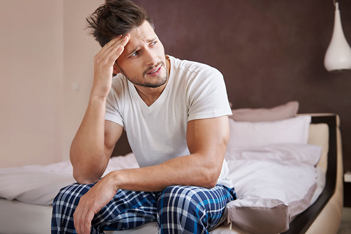 Man sitting on bed looking stressed and frustrated, wearing a white t-shirt and blue plaid pants in bedroom. Man sitting on bed looking stressed and frustrated, wearing a white t-shirt and blue plaid pants in bedroom.