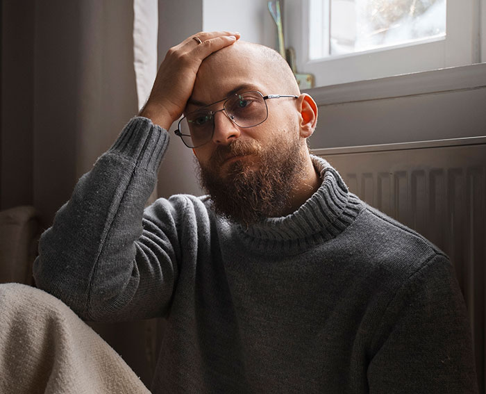 Man sitting indoors looking frustrated and stressed, wearing glasses and a gray sweater near a window. Man sitting indoors looking frustrated and stressed, wearing glasses and a gray sweater near a window.