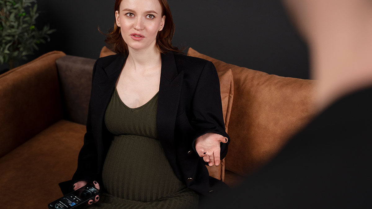 Pregnant woman in a green dress and black blazer sitting on a couch, speaking with a man during a tense moment.
