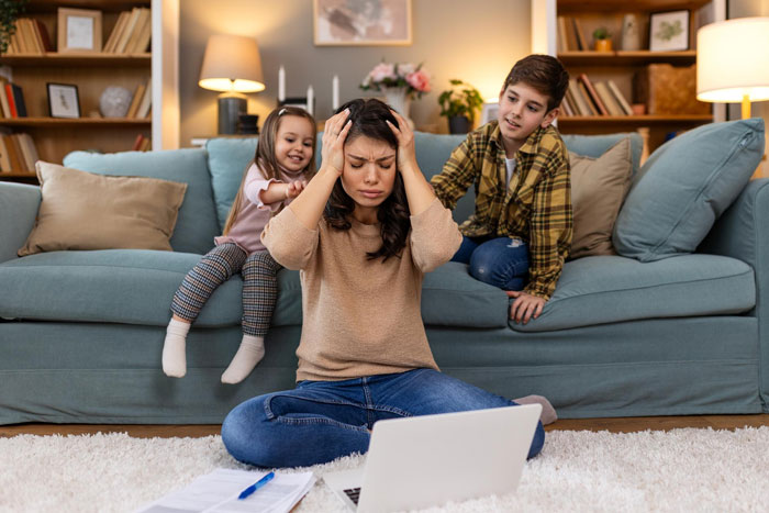 Stressed mom of 3 sitting on floor with laptop while children play on couch behind her in cozy living room. Stressed mom of 3 sitting on floor with laptop while children play on couch behind her in cozy living room.