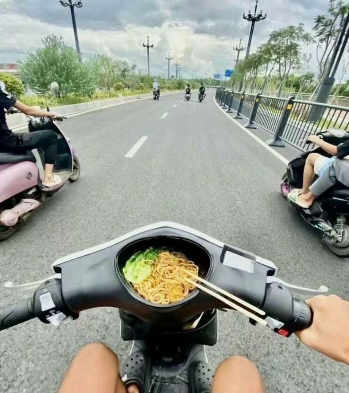 View from scooter rider showing noodles and chopsticks in scooter dashboard on a city street, capturing pleasures of the occult humor.