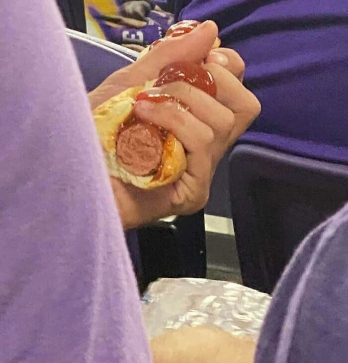 Hand holding a hot dog with ketchup, captured in an out-of-context moment fitting the pleasures of the occult theme.