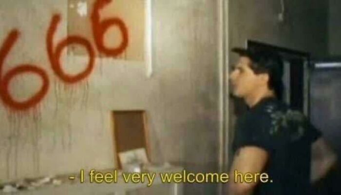 Man standing in a dimly lit room with graffiti of 666 on the wall, captioned feeling very welcome in an occult setting.