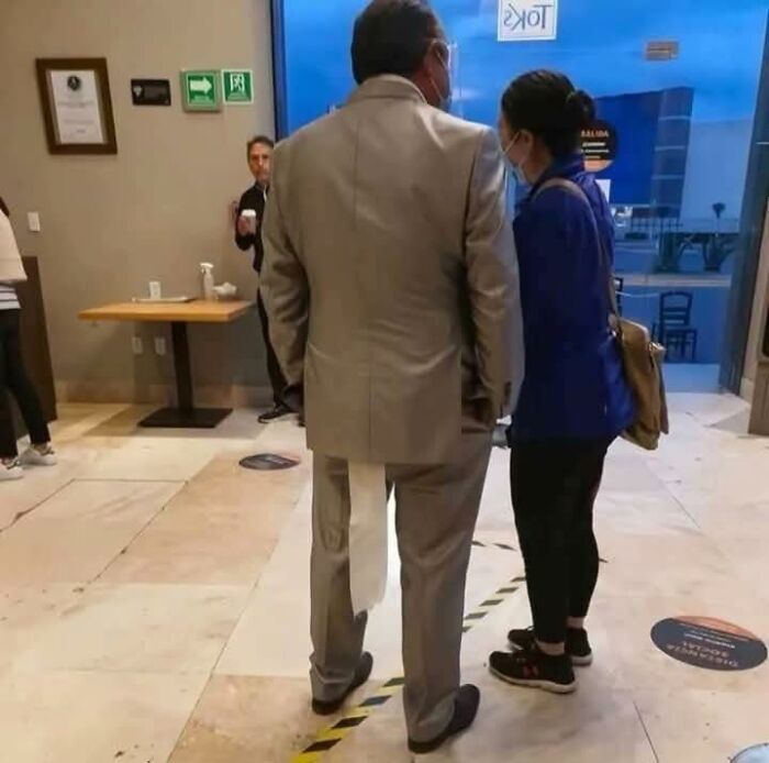 Man in a suit with a long strip of toilet paper stuck to his pants, standing indoors near a woman wearing a blue jacket.