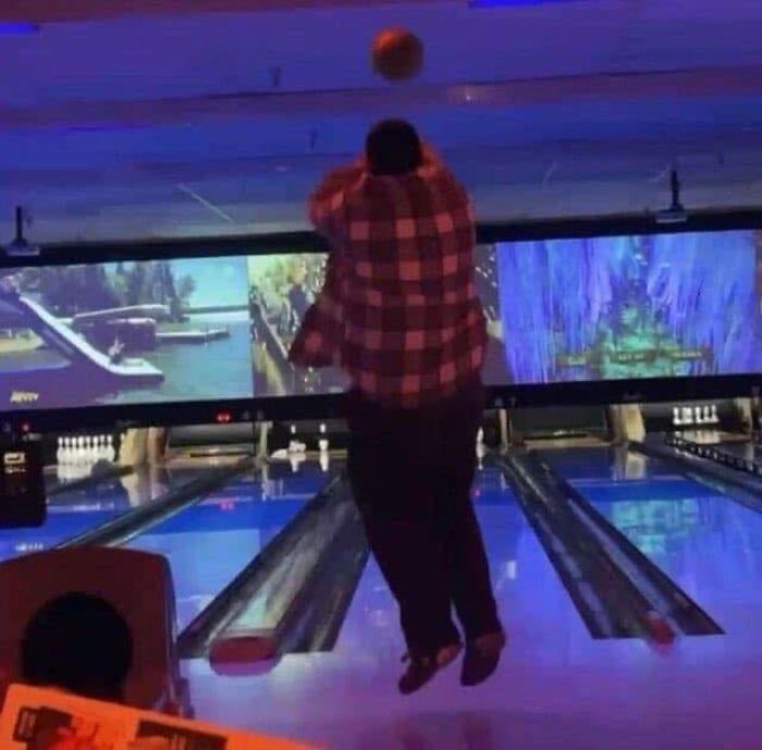 Person in a red plaid shirt jumping while bowling indoors with scenic visuals on screens above the lanes, pleasures of the occult theme.
